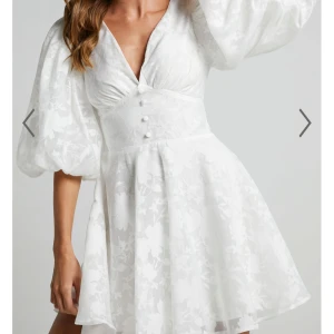 Vit studenttklänning med puffärmar från Showpo - Superfin vit kortklänning från Showpo med v-ringning, markerad midja och stora puffärmar. Klänningen har subtilt blommigt mönster i tyget och knappar framtill. Perfekt för sommarens tillfällen!