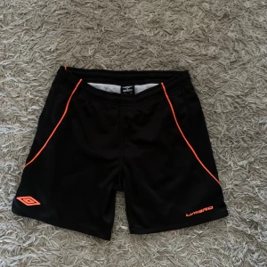 Svarta badshorts från Umbro med orange detaljer - Svarta träningsshorts från Umbro med orangea detaljer längs sidorna och logga framtill. Shortsen har resår i midjan och är tillverkade i ett lätt och svalt material, perfekt för sport och aktivitet.