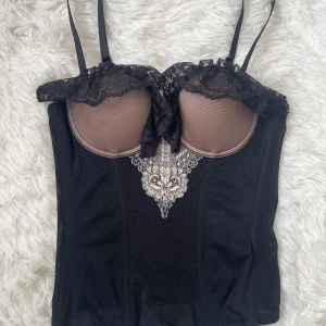 Svart korsettopp med spetsdetaljer - Säljer en svart korsettopp med tunna axelband och vacker spets upptill samt broderad spetsdetalj framtill. Toppen har vadderade kupor i beige och knäppning bak. Baksidan är mer genomskinlig än framsidan (se bild 4 och 5). Passar byst 78-80cm, underbyst 65-70cm, midjan 58-62cm.