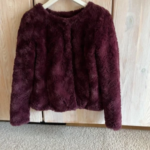 Vinröd pälsjacka från Vero Moda - Mysig vinröd pälsjacka från Vero Moda med lång ärm och rund halsringning. Jackan har knäppning framtill och är fodrad på insidan. Perfekt för att addera lite extra stil till din outfit.