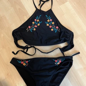 Svart bikini med broderade blommor - Svart bikini med färgglada broderade blommor på både överdel och underdel. Överdelen har hög hals och knyt i sidan, medan underdelen har matchande broderi framtill. Perfekt för sommaren!