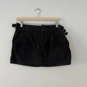 Svart minikjol  - Svart minikjol med två knappar framtill, snedställda fickor och bältesdetaljer i sidorna. Kjolen har även fickor med lock baktill och en rak passform. 