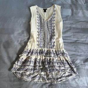 Vit och blå ärmlös klänning med mönster från H&M - En så drömmig klänning från H&M i vitt med blått etnoinspirerat mönster. Klänningen har v-ringning och volangdetaljer 🪸🐚 Endast använd ett fåtal gånger. Fint skick! 