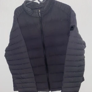 Svart pufferjacka från Moose Knuckles - Säljer en svart pufferjacka från Moose Knuckles med diskret logga på ärmen och camo-detaljerad inneretikett. Jackan har dragkedja framtill och quiltad design som ger en modern look. Perfekt för mysiga dagar.