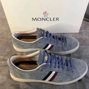 Grå mockasneakers från Moncler - Säljer ett par grå sneakers från Moncler i mocka med vita sulor och blå snörning. Skorna har Monclers klassiska ränder i rött, vitt och blått på sidan samt rund tå. Perfekta för dig som gillar stilrena och exklusiva sneakers. Skick 8/10, perfekta till sommaren, box ingår. Skriv om ni undrar något. Pris kan diskuteras vid snabb affär.