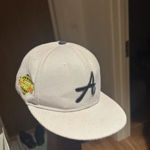 Ljusgrå keps med svart A-logga - Snygg ljusgrå keps med svart broderad A-logga framtill och ett färgglatt märke på sidan. Platt skärm och klassisk passform. Perfekt accessoar för en sportig look.