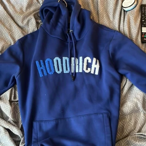 Blå hoodie från Hoodrich - Säljer en blå hoodie från Hoodrich med broderad logga i olika blå och vita nyanser på bröstet. Tröjan har huva med dragsko och en stor magficka. Perfekt för dig som gillar streetwear och vill ha något bekvämt och snyggt.