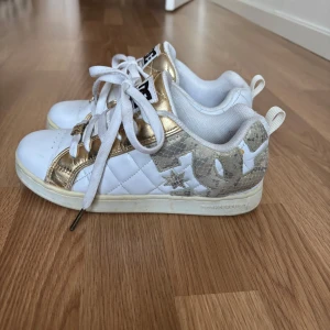 DC Shoes sneakers i vitt och guld - Säljer ett par DC Shoes sneakers i vitt med metallicguld-detaljer och stora DC-loggor på sidorna. Skorna har snörning, quiltad textur på sidan och platt sula. Perfekta för dig som gillar streetstyle och vill sticka ut.