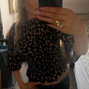 Svart och beige mönstrad blus med hög hals - Trendig blus i svart med beige mönster och hög hals. Modellen har trekvartsärm och smockad midja som ger en snygg siluett. Perfekt till jeans eller kjol för en cool look.