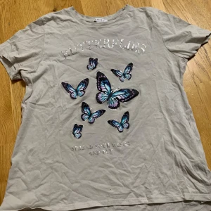 Beige t-shirt med fjärilar och text - Säljer en beige t-shirt med glansig text 'BUTTERFLIES' och flera tryckta fjärilar i blått, lila och svart på framsidan. T-shirten har rund hals och korta ärmar. Perfekt för dig som gillar färgglada detaljer!