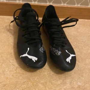 Svarta Puma Future fotbollsskor - Säljer ett par svarta Puma Future fotbollsskor med vita detaljer och logga. Skorna har snörning och är designade för att ge bra grepp på planen. Tyvärr så kommer skorna utan låda fast dom är i jätte bra skick. Dom är heller inte använda. Perfekta för dig som vill ha stilrena och funktionella fotbollsskor.