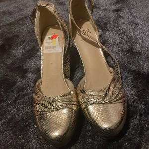 Guldiga klackskor från VOX - Säljer ett par glansiga guldiga klackskor från VOX med ormskinnsmönster och bred klack. Skorna har en rem runt vristen med spänne och rund tå. Tillverkade i syntetmaterial och märkta som vegan shoes.