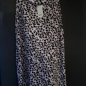 Ljuslila leopardmönstrad långkjol - Säljer en långklänning i ljuslila med svart leopardmönster. Klänningen har en lös passform och är ärmlös med snygg drapering vid halsen. Perfekt för dig som vill sticka ut med ett trendigt mönster.