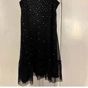 Svart prickig klänning med volang - Säljer en svart klänning med tunna axelband och små färgglada prickar över hela tyget. Klänningen har volangdetalj nertill och är lätt och luftig, perfekt för varma dagar eller festliga tillfällen.