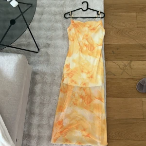 Gul och orange mönstrad långklänning med smala axelband - Säljer en somrig långklänning med smala axelband och vackert abstrakt mönster i gula och orange toner. Klänningen har en figurnära passform och är tillverkad i ett lätt, syntetiskt material. Perfekt för varma dagar!
