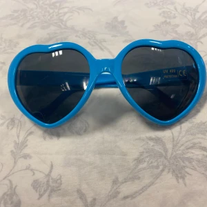 Hjärt solglasögon - Två par hjärtformade solglasögon, ett par med blå bågar och ett par med vita bågar. Båda har mörka linser och en lekfull design som sticker ut. Perfekta för att ge din stil en rolig touch. 75kr/par , helt ny 