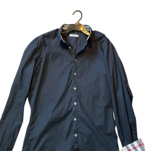Svart skjorta burberry  - Snygg mörkblå skjorta från burberry med klassisk krage och knappar framtill. Skjortan har långa ärmar och en stilren design, perfekt för dig som gillar en clean och tidlös look. Manschetterna har ett diskret rutigt tyg på insidan för en extra detalj.