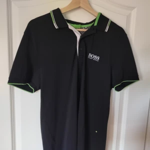 Svart pikétröja från Hugo BOSS - Säljer en svart pikétröja från BOSS med vit logga på bröstet och nacken. Tröjan har gröna detaljer på krage och ärmslut.