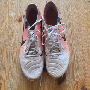 Nike fotbollsskor i vitt och rosa - Säljer ett par Nike fotbollsskor i vitt och ljusrosa med svart swoosh. Skorna har snörning och rund tå, samt platt sula med dobbar för bra grepp på planen. Perfekta för dig som vill sticka ut på fotbollsplanen! Lite slitna i framkant och använda men är i bra skick! Storlek 44, nypris 2799! Hör av dig vid frågor🤗