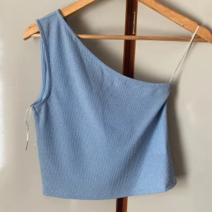 Ljusblå one shoulder topp från Pieces - Säljer en ljusblå ribbad one shoulder topp från Pieces. Toppen har en asymmetrisk design med en axel och är tillverkad i ett mjukt, stretchigt material. Perfekt för att skapa en trendig look. Endast använd 1 gång