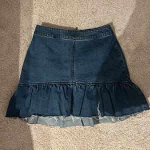 Mörkblå jeanskjol med volang - Säljer en mörkblå jeanskjol med volang nertill och rå kant. Kjolen har dragkedja bak och markerad söm framtill. Super fin!!