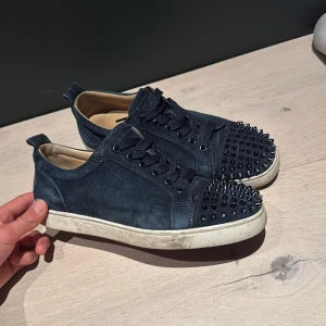 Christian Louboutin skor - Säljer ett par svarta sneakers i mocka med svarta nitar på tån. Skorna har snörning och rund tå, samt vit sula. Insidan är ljus och det står 'Paris' tryckt i skon. Perfekta för dig som vill sticka ut lite extra. Dessa skor är extremt fina och sällsynta, Vissa spikar är blekta på toppen på grund av att de har slitits och att färgen har åkt av och en av spikarna är helt blekt fast det är inget man märker. box, dustbag medföljer. Pris är diskutabelt!