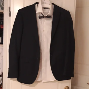 Mörkblå kostym från Dressmann - Klassisk mörkblå kostym från Dressmann i storlek 46 regular, slim fit, byxor storlek 32/30. Passar smal kille cirka 176-180 cm lång. Killen på bilden är 183cm och väger ca 74 kg och då är kostymen lite för liten. Använd en gång. Perfekt för bal eller student.. Skjorta och fluga kan ingå om så önskas.