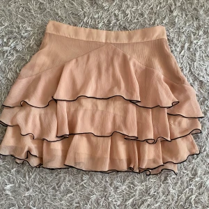 Beige volangkjol från Zara - Supersöt beige kjol från Zara med flera lager volanger och svarta kanter. Kjolen har en kort modell och är tillverkad i ett lätt, plisserat tyg som ger ett luftigt intryck. Perfekt för dig som gillar romantiska och trendiga plagg. Jätteskönt material. 