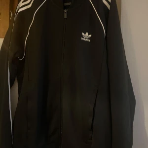Svart Adidas track jacket med vita ränder - Klassisk svart Adidas track jacket med vita ränder längs axlarna och ärmarna. Storlek M passar perfekt, använd ett fåtal gånger. Nypris 600kr