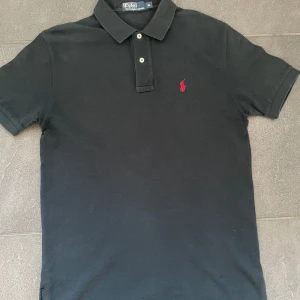 Ralph Lauren Pike - Klassisk svart pikétröja från Polo Ralph Lauren med röd broderad logga på bröstet. Tröjan har krage, knappar framtill och korta ärmar.                      Storlek: M.                                                                           Skick: Mycket bra😊