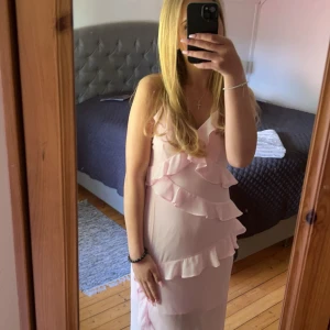 Ljusrosa långklänning med volanger - Säljer en ljusrosa långklänning med smala axelband och v-ringning. Klänningen har flera volangdetaljer framtill och längs kjolen, vilket ger en romantisk och feminin känsla. Perfekt för sommarens tillfällen. Endast använd 1 gång, den är slutsåld och svårt att hitta💕