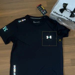 Under Armour Reflective Set - Säljer ett par svarta träningsshorts och tränings t-shirt från Under Armour med elastisk midja och färgglada reflektiva detaljer på sidorna. Shortsen har logga på vänster ben och är perfekta för träning eller sport.