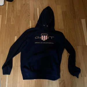 Mörkblå hoodie från GANT - Säljer en mörkblå hoodie från GANT med tryckt logga och texten 'American Sportswear' på bröstet. Tröjan har huva med dragsko och klassisk känguruficka framtill. Perfekt för en sportig och avslappnad stil.