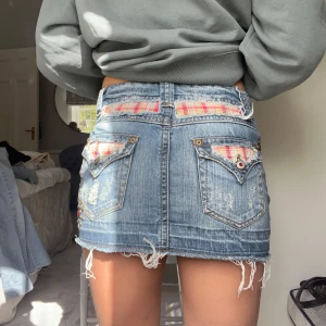 Jeanskjol med rutiga detaljer - Säljer en kort jeanskjol med slitna kanter och rutiga detaljer vid fickorna. Kjolen har två knappar framtill och dekorativa nitar på ena fickan. Perfekt för dig som gillar en edgy och trendig stil.