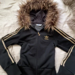 adidas fur hoodie - Svart Adidas jacka med guldiga ränder på ärmarna och dragkedja framtill. Huva med fuskpäls som är påsydd själv. Fuskpälsen är från en jacka som nästan aldrig använts. Storlek M men passar definitivt S 