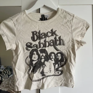 Vit Black Sabbath t-shirt från H&M Divided - Cool vit t-shirt från H&M Divided med Black Sabbath-tryck och porträtt av bandmedlemmarna på framsidan. T-shirten har korta ärmar och en croppad passform, perfekt för dig som gillar rockstil, så snygg!💘😌