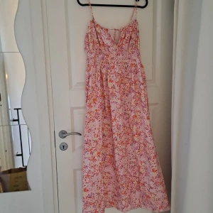 Blommig sommarklänning med smock - Säljer en söt blommig klänning med smockdetaljer och tunna axelband. Klänningen har en härlig mix av rosa, lila och orange blommor på en ljus bakgrund. Perfekt för varma sommardagar!