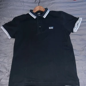 Svart pikétröja från Hugo Boss - Snygg svart pikétröja från Hugo Boss med vit broderad logga på bröstet. Tröjan har en klassisk krage med vita och grå ränder samt knappar framtill. Perfekt för en stilren look. (Pris kan diskuteras)