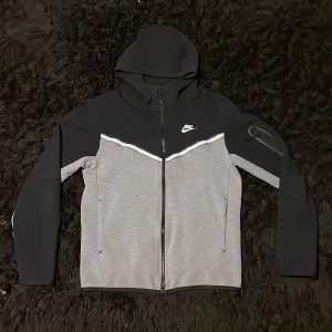  Nike tech fleece - Säljer en snygg grå och svart hoodie från Nike med dragkedja och huva. Jackan har en sportig design med vita detaljer och är perfekt för en casual look. Den har långa ärmar och är tillverkad i ett mjukt material. Hör av dig om du har några funderingar.
