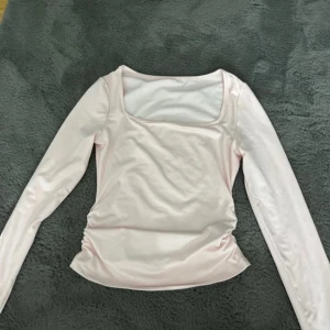 Ljusrosa långärmad topp - Säljer en söt ljusrosa långärmad topp med rynkade detaljer på sidorna. Toppen har en fyrkantig halsringning och är gjord i ett mjukt material. Perfekt för en stilren look! I storlek XS/S, aldrig använd är i ny skick.