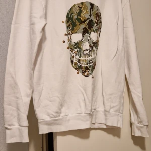 Vit sweatshirt med dödskallemotiv(oanvänd) - Säljer en vit sweatshirt med ett stort dödskallemotiv i camouflage och paljetter på framsidan. Tröjan har långa ärmar och ribbade muddar vid ärmslut och nederkant. Perfekt för en cool och avslappnad stil.