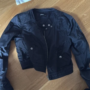 Svart jacka från Zoul - Snygg svart jacka från Zoul med dragkedja och knappar. Jackan har quiltade detaljer på axlarna och en croppad passform. Perfekt för en stilren look.