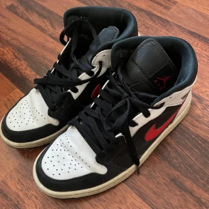 Nike Air Jordans i svart, vitt och rött - Snygga Nike Air Jordans i klassisk färgkombination av svart, vitt och rött. Skorna har en hög modell med snörning och den ikoniska swoosh-loggan i rött på sidan. Perfekta för både stil och komfort.