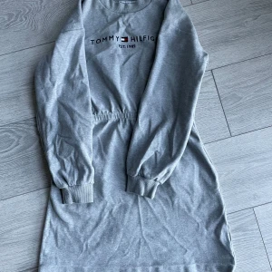 Grå klänning från Tommy Hilfiger - Snygg grå klänning från Tommy Hilfiger med tryck framtill. Klänningen har långa ärmar och en bekväm passform med resår i midjan. Perfekt för en avslappnad stil.