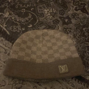 Rutig mössa  - Snygg och stilren mössa  med ett klassiskt rutigt mönster. Mössan är i en neutral färgton och har en uppvikt kant med en diskret logotyp. Perfekt för att hålla värmen med stil.