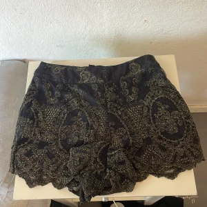 Svarta shorts med glittrigt mönster - Snygga svarta shorts med ett glittrigt mönster som ger en festlig känsla. Shortsen har en elegant design med spetsdetaljer längs kanterna. Perfekta för att sticka ut på nästa fest! Sååååå snygga❤️