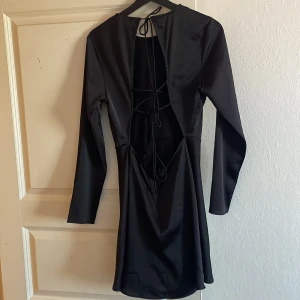 Svart klänning från Gina Tricot - Snygg svart klänning från Gina Tricot med långärmad design och knytning i ryggen. Perfekt för en elegant look med sin stilrena och klassiska stil. Klänningen har en smickrande passform och är tillverkad i ett mjukt material.