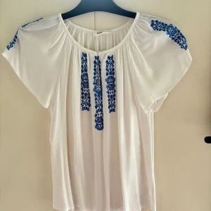 Vit blus med blå broderier från KappAhl - Säljer en söt vit blus från KappAhl med vackra blå broderier på bröstet och axlarna. Blusen har korta ärmar och en luftig passform, perfekt för varma dagar. Passar både till jeans och kjol! Passar som en XS!
