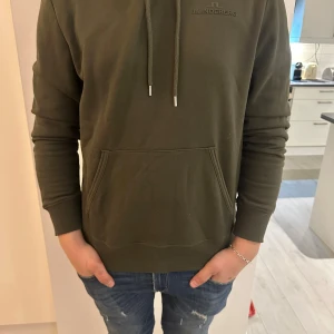 Olivgrön hoodie från J.Lindeberg - Säljer en stilren olivgrön hoodie från J.Lindeberg. Tröjan har en klassisk design med dragsko i huvan och en praktisk magficka. Köpt på design only i somras. Storlek M, jag är 181 och väger ca 75kg.  Nypris 1400kr :).