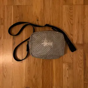 Säljer en stilren grå axelväska från Stussy med ett diskret rutmönster och justerbar axelrem. Perfekt för att bära med sig det nödvändigaste på ett snyggt sätt.